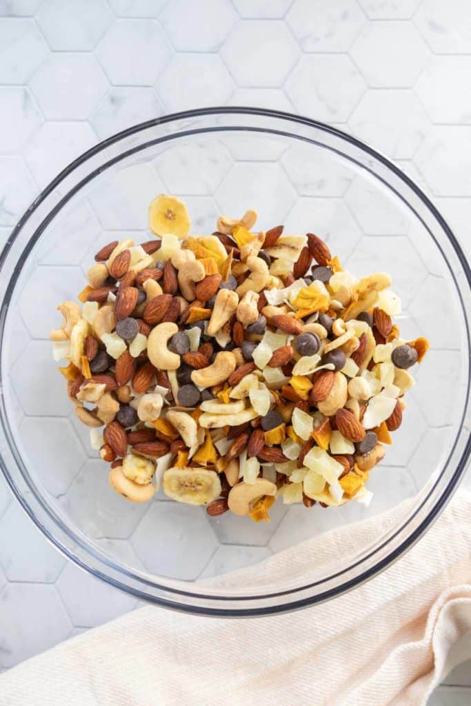 A glass bowl filled with a mix of nuts, dried fruits, banana chips, and chocolate chips on a white hexagonal tile surface.