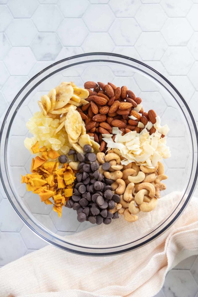 A glass bowl containing almonds, cashews, chocolate chips, dried banana chips, dried pineapple, coconut flakes, and dried mango on a white hexagon-tile surface.