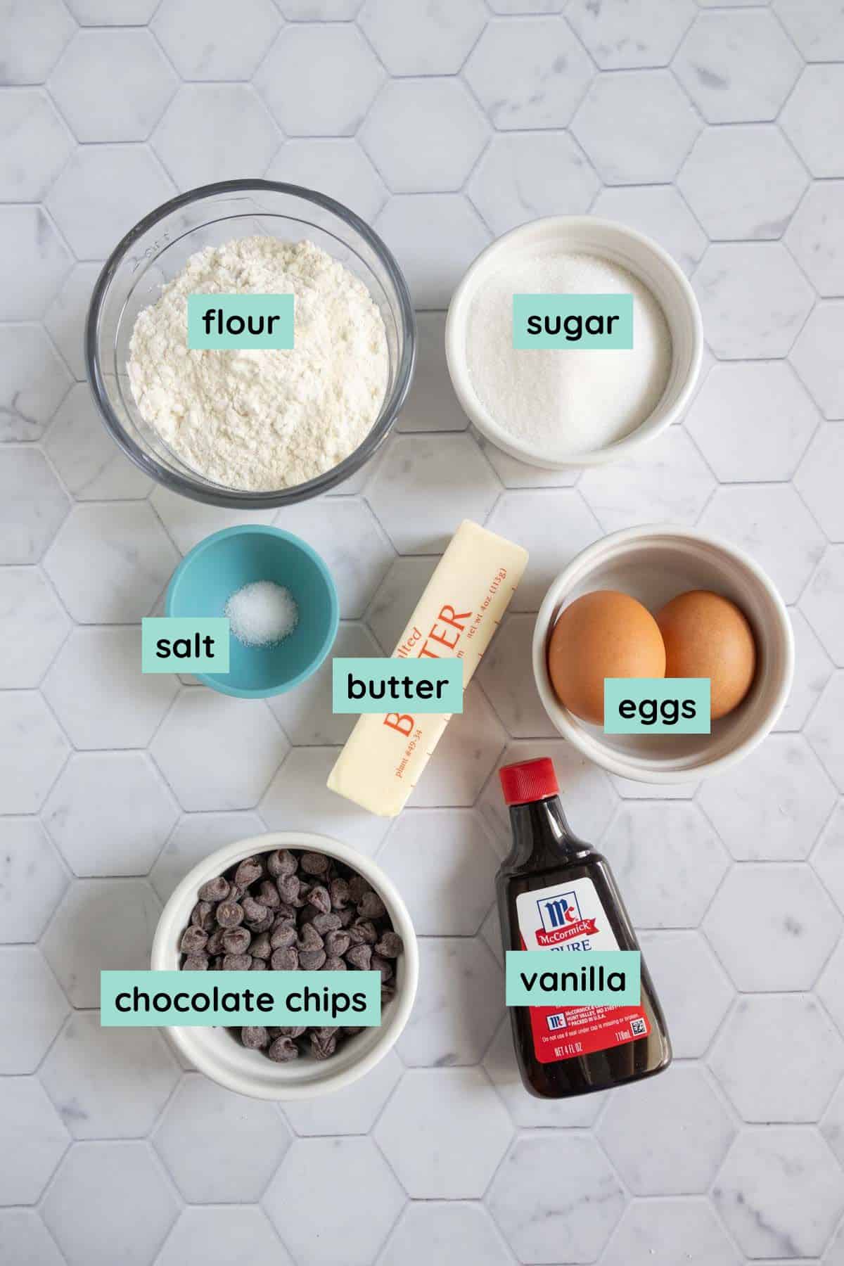 Overhead view of labeled baking ingredients: flour, sugar, salt, butter, eggs, chocolate chips, and vanilla arranged on a white hexagonal tile surface.