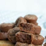 A stack of chocolate muffins, with one muffin on top cut in half to show a moist, dense interior. Other muffins are arranged in the background on a wooden board.