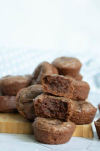 A stack of chocolate muffins, with one muffin on top cut in half to show a moist, dense interior. Other muffins are arranged in the background on a wooden board.