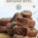 A stack of brownie bites with one cut in half to show the moist, dense interior, placed on a wooden board. Text reads "Brownie Bites" and "happysnackcidents.com".