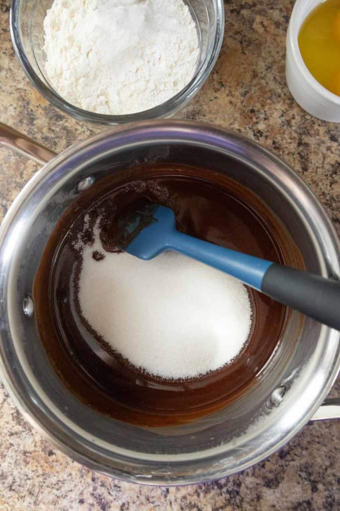A saucepan with melted chocolate and sugar being mixed with a blue spatula, with a bowl of flour and a bowl of eggs nearby on a countertop.