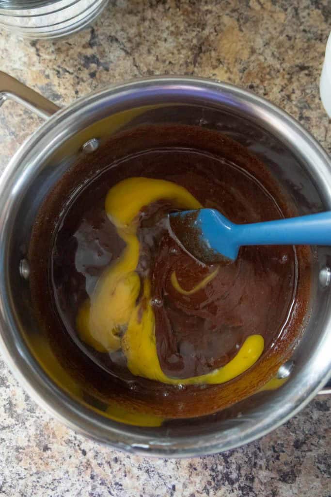 A blue spatula stirs egg yolks into a chocolate mixture in a stainless steel pot on a speckled countertop.