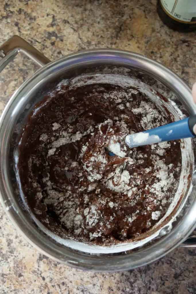 A metal mixing bowl filled with chocolate brownie batter being mixed with a blue spatula, sitting on a speckled countertop.