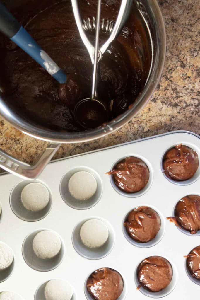 A mixing bowl with chocolate batter and a scoop next to a muffin tin, some wells filled with batter, on a kitchen counter.