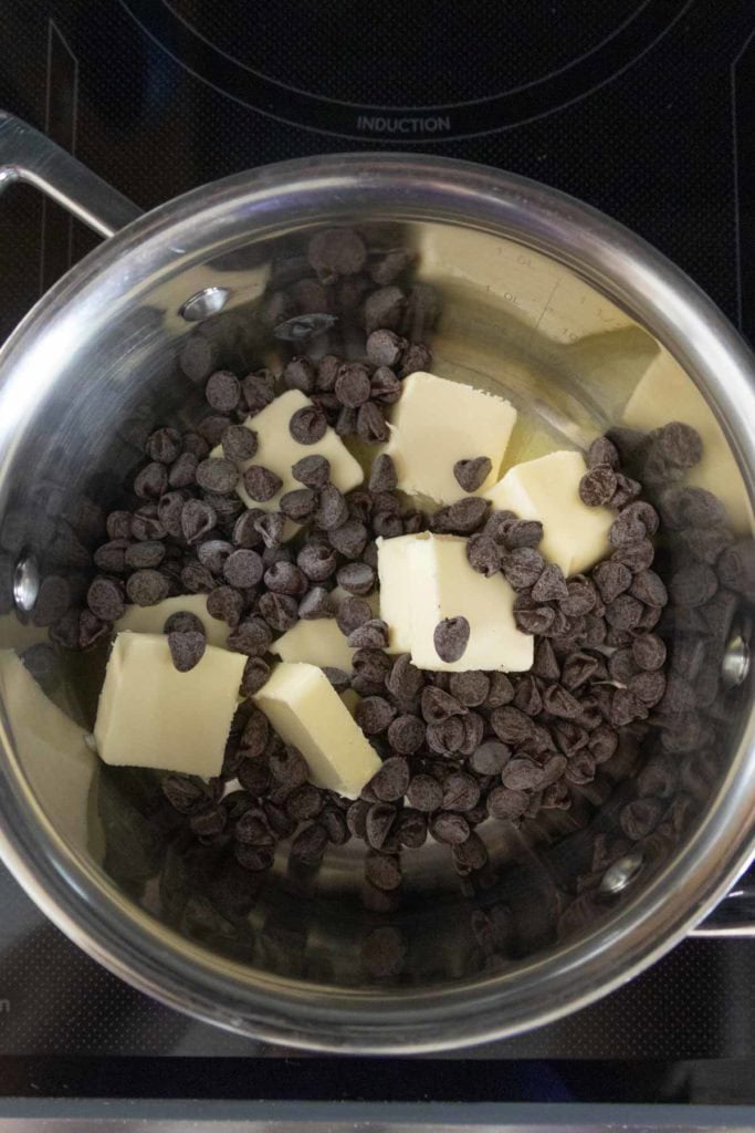 A stainless steel saucepan on a stovetop containing chunks of butter and semi-sweet chocolate chips before melting.