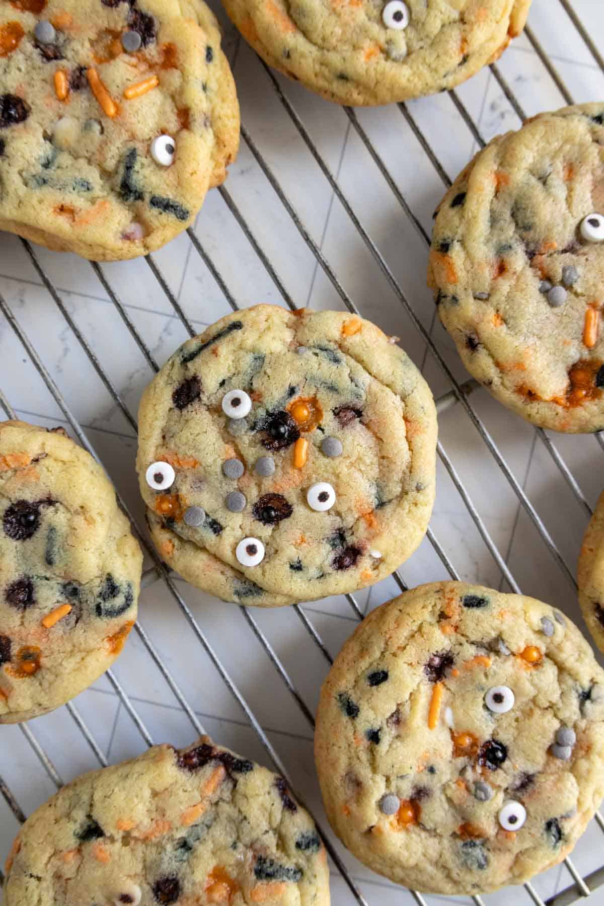 Cookies with colorful sprinkles and candy eyes rest on a wire cooling rack.
