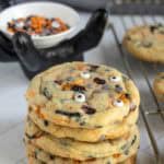 A stack of Halloween-themed cookies with candy eyes and colored candy pieces, next to a cooling rack and a bowl of festive sprinkles.