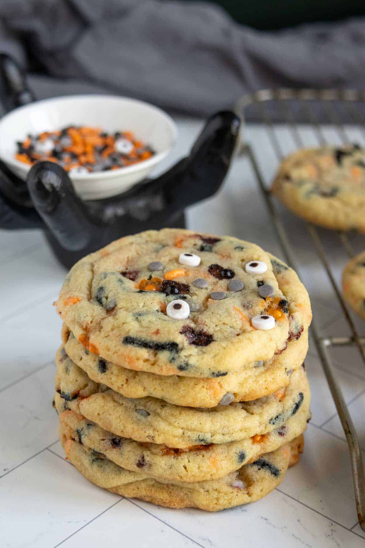 A stack of Halloween-themed cookies with candy eyes and colored candy pieces, next to a cooling rack and a bowl of festive sprinkles.