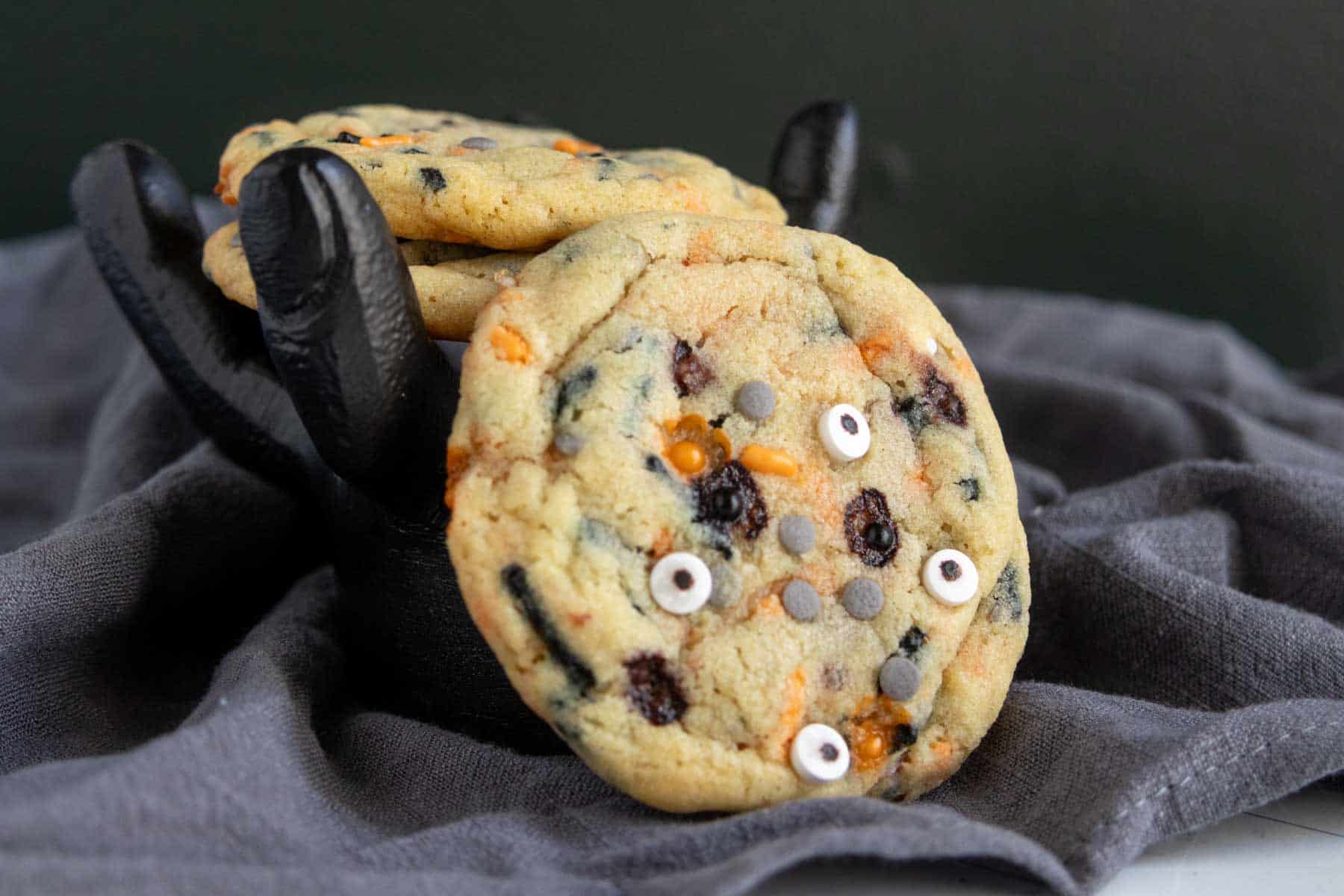 A stack of cookies with chocolate chips and candy eyes rests on a black decorative hand atop a dark gray cloth.