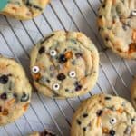 Round Halloween-themed cookies with chocolate chips, orange sprinkles, and candy eyes on a cooling rack. Text at the bottom reads "HALLOWEEN COOKIES happysnackcidents.com".