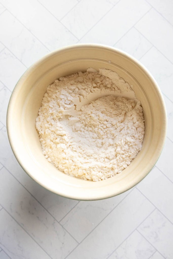 A beige mixing bowl filled with flour sits on a white tiled surface.