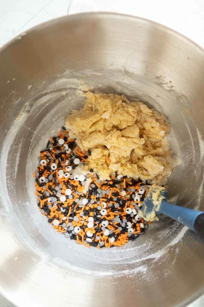 A mixing bowl with cookie dough and black, orange, and white Halloween-themed sprinkles being stirred with a blue spatula.