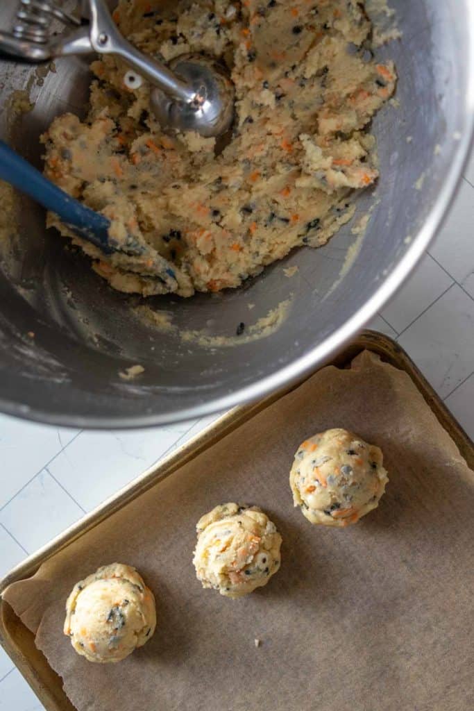 A metal mixing bowl with cookie dough and a scoop, next to a baking tray lined with parchment paper holding three scoops of dough.