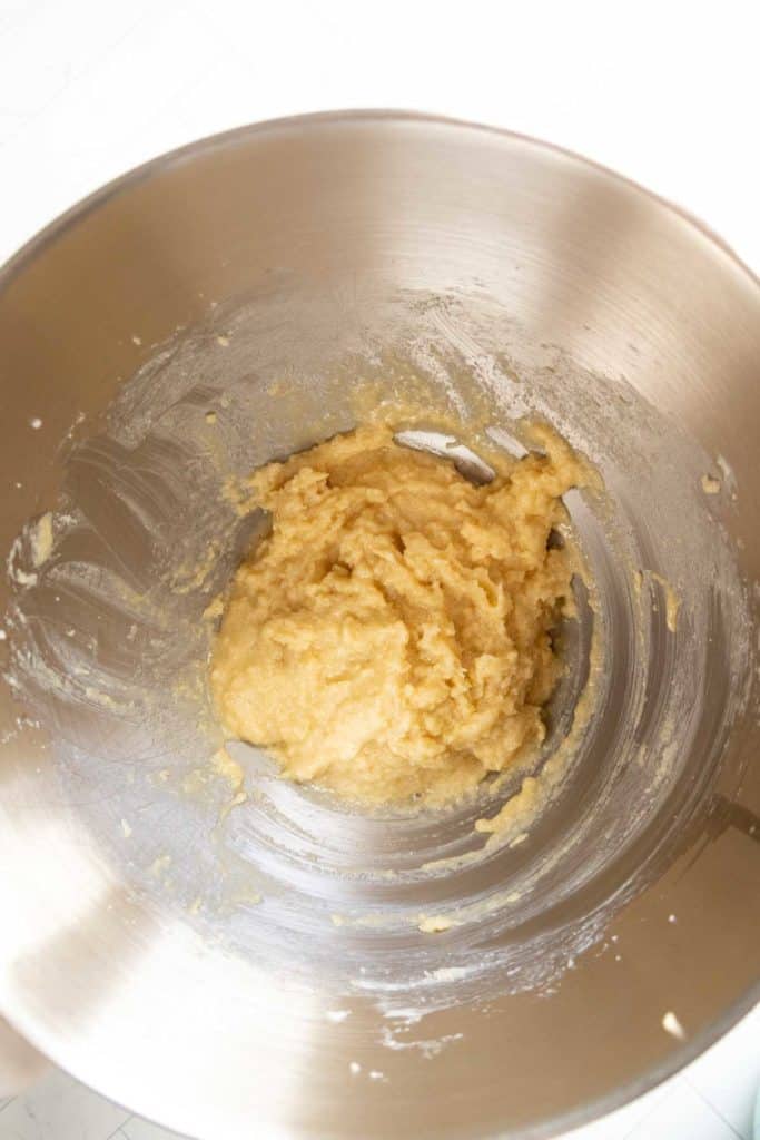 A stainless steel mixing bowl containing a partially mixed, light-colored dough or batter sits on a white surface.