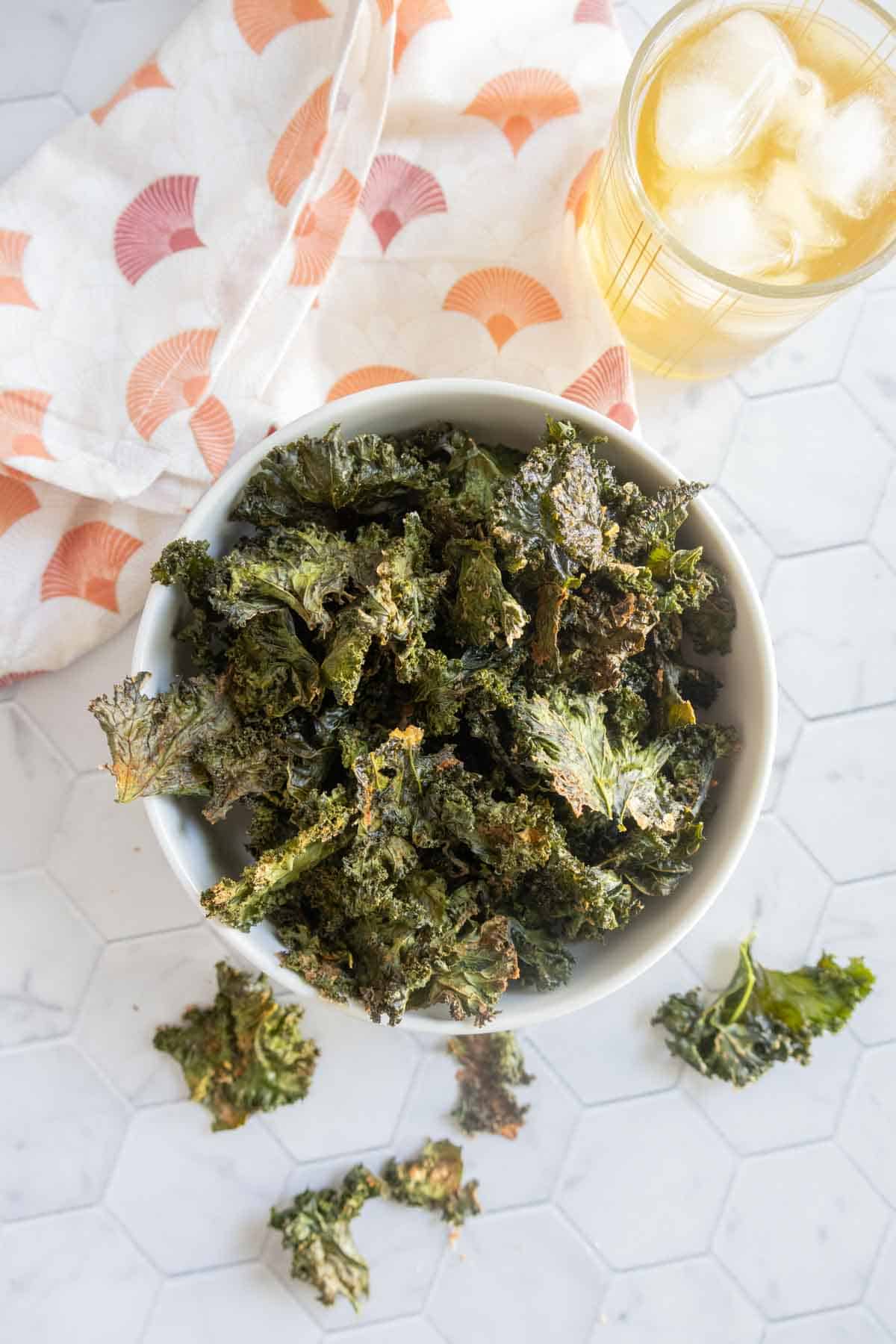 A bowl of crispy kale chips on a white hexagonal tile surface, with a patterned napkin and a glass of iced drink nearby.