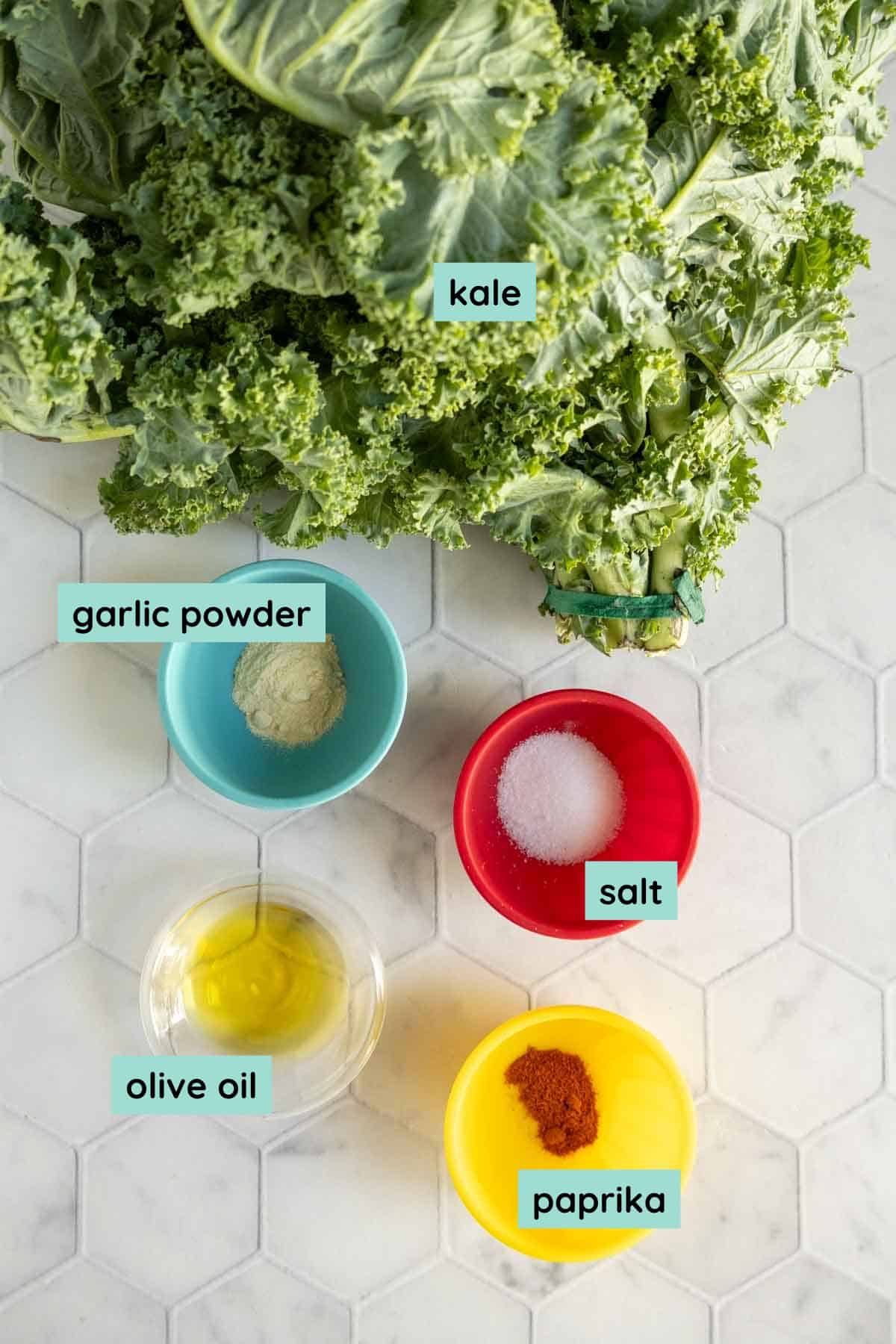 Fresh kale leaves with labeled small bowls containing garlic powder, salt, olive oil, and paprika are arranged on a white hexagonal tile surface.