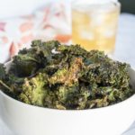 A white bowl filled with baked kale chips sits on a white surface, with a glass of a light-colored drink in the background.