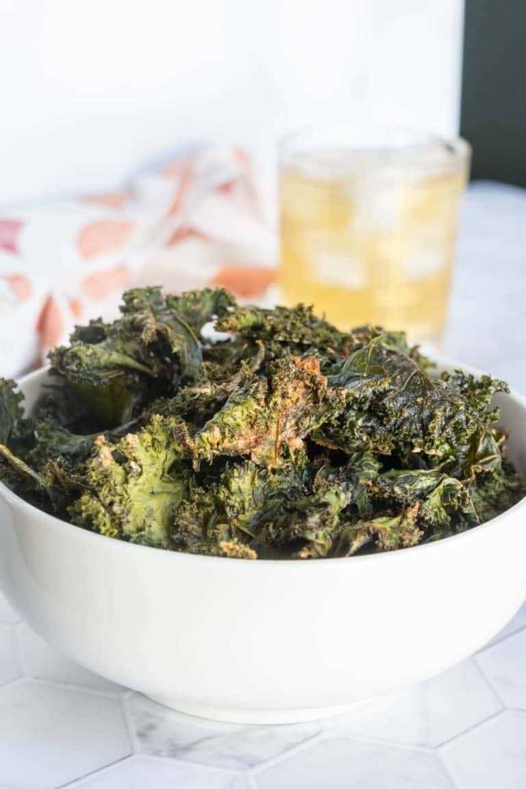 A white bowl filled with baked kale chips sits on a white surface, with a glass of a light-colored drink in the background.