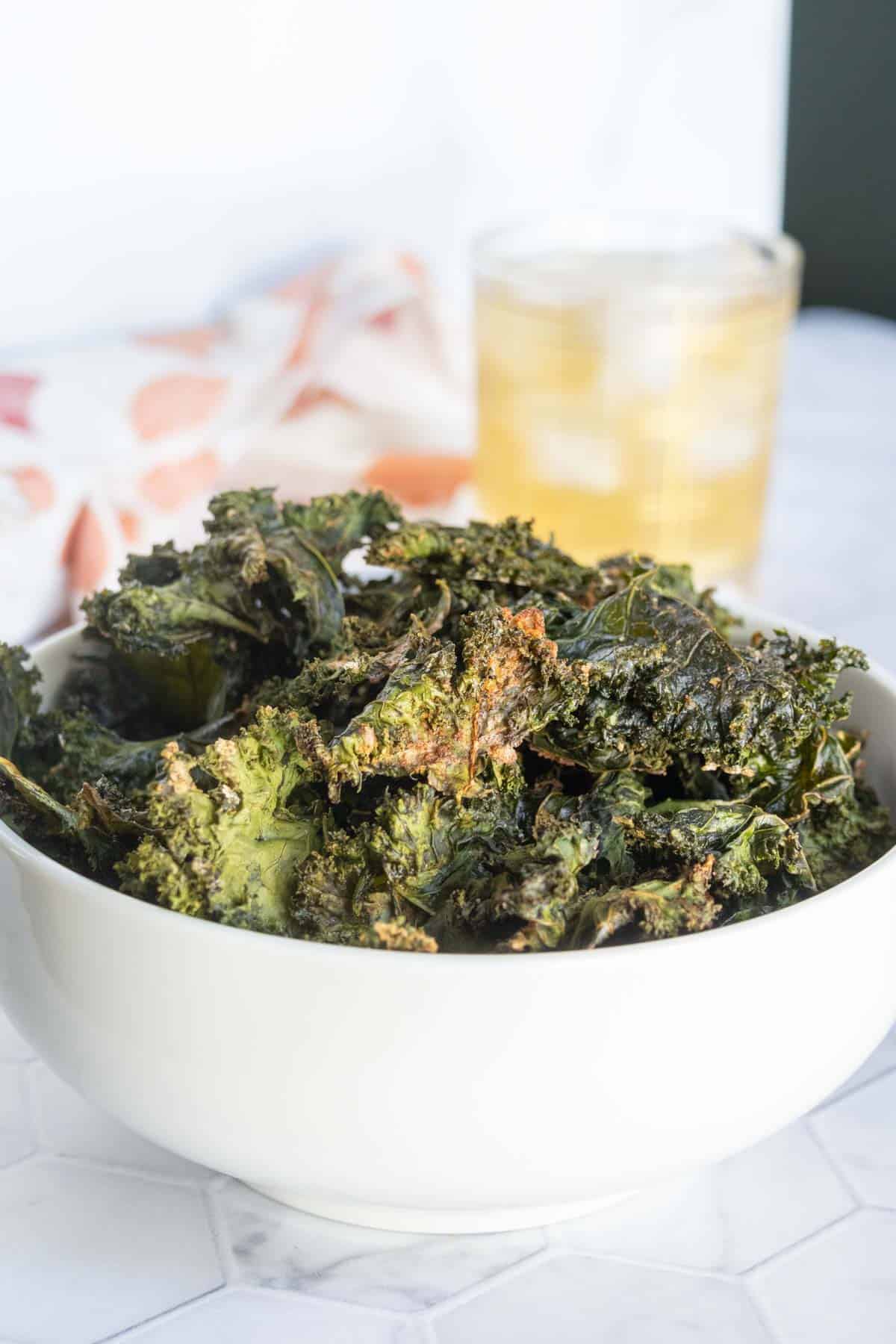 A white bowl filled with baked kale chips sits on a white surface, with a glass of a light-colored drink in the background.