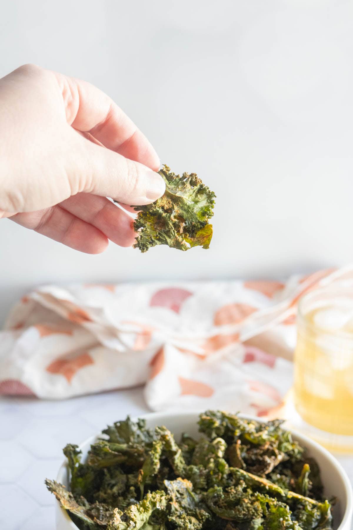 A hand holds a piece of kale chip above a bowl filled with more kale chips, with a napkin and drink in the background.