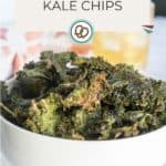A white bowl filled with crispy kale chips sits on a table with a drink in the background. The image includes the text "Kale Chips" and "happysnackcidents.com".