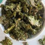 A bowl of baked kale chips sits on a white surface, with a few chips scattered around the bowl. Text at the bottom reads "Kale Chips" and "happysnackcidents.com".