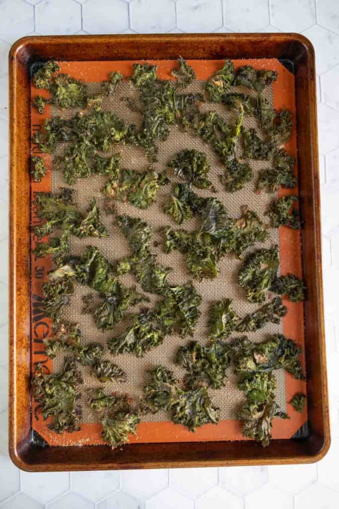 A baking sheet covered with a nonstick mat holds scattered, baked kale chips on a marble countertop.