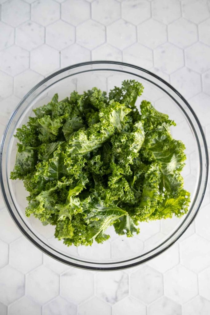 A clear glass bowl filled with fresh, chopped kale leaves sits on a white, hexagon-tiled surface.