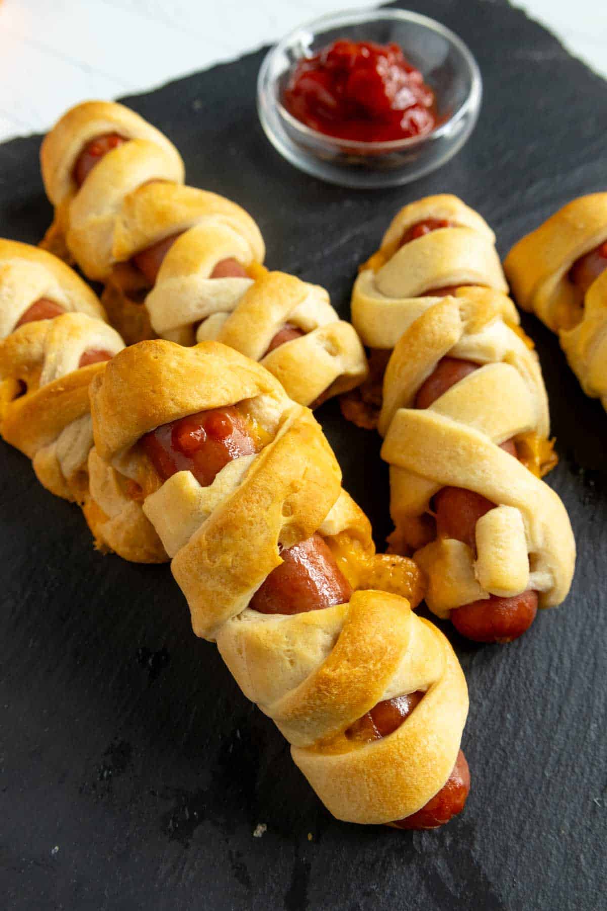 Hot dogs wrapped in baked crescent roll dough are arranged on a dark serving board, with a small dish of ketchup in the background.