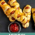 Three hot dogs wrapped in crescent dough to resemble mummies are served with a small bowl of ketchup on a dark surface. Text at the bottom reads "Mummy Hot Dogs, happysnackcidents.com.