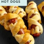 Several hot dogs are wrapped in crescent roll dough to resemble mummies, with a small bowl of ketchup in the background on a dark serving board.
