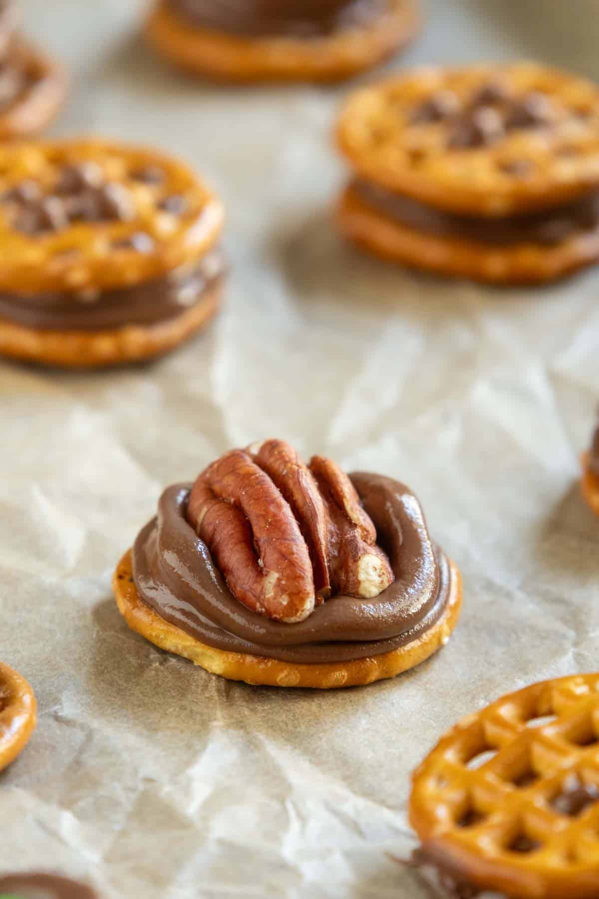 A pretzel round topped with a swirl of chocolate and a pecan half, set on parchment paper with similar treats in the background.