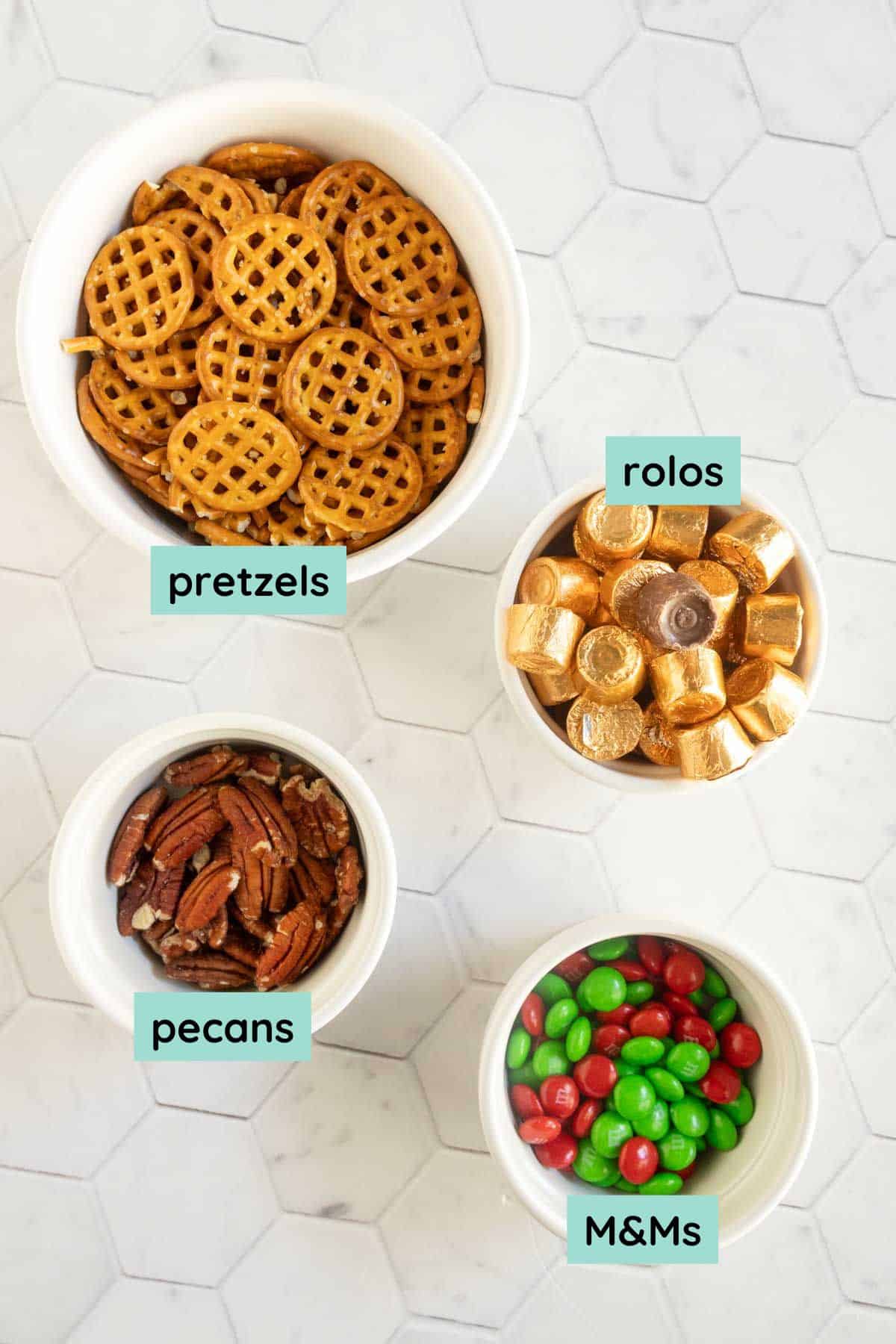 Four bowls on a white surface, containing pretzels, Rolos chocolates, pecans, and red and green M&Ms, each labeled with text.
