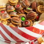 A festive tin filled with pretzel-based treats topped with chocolate, pecans, and red or green candies, surrounded by gold-wrapped chocolates and a red ribbon.