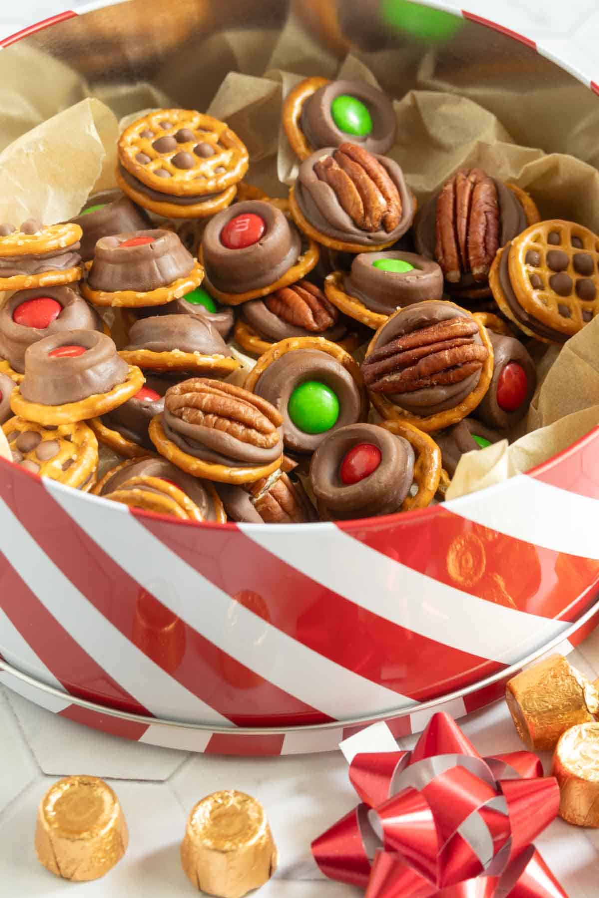 A festive tin filled with pretzel-based treats topped with chocolate, pecans, and red or green candies, surrounded by gold-wrapped chocolates and a red ribbon.