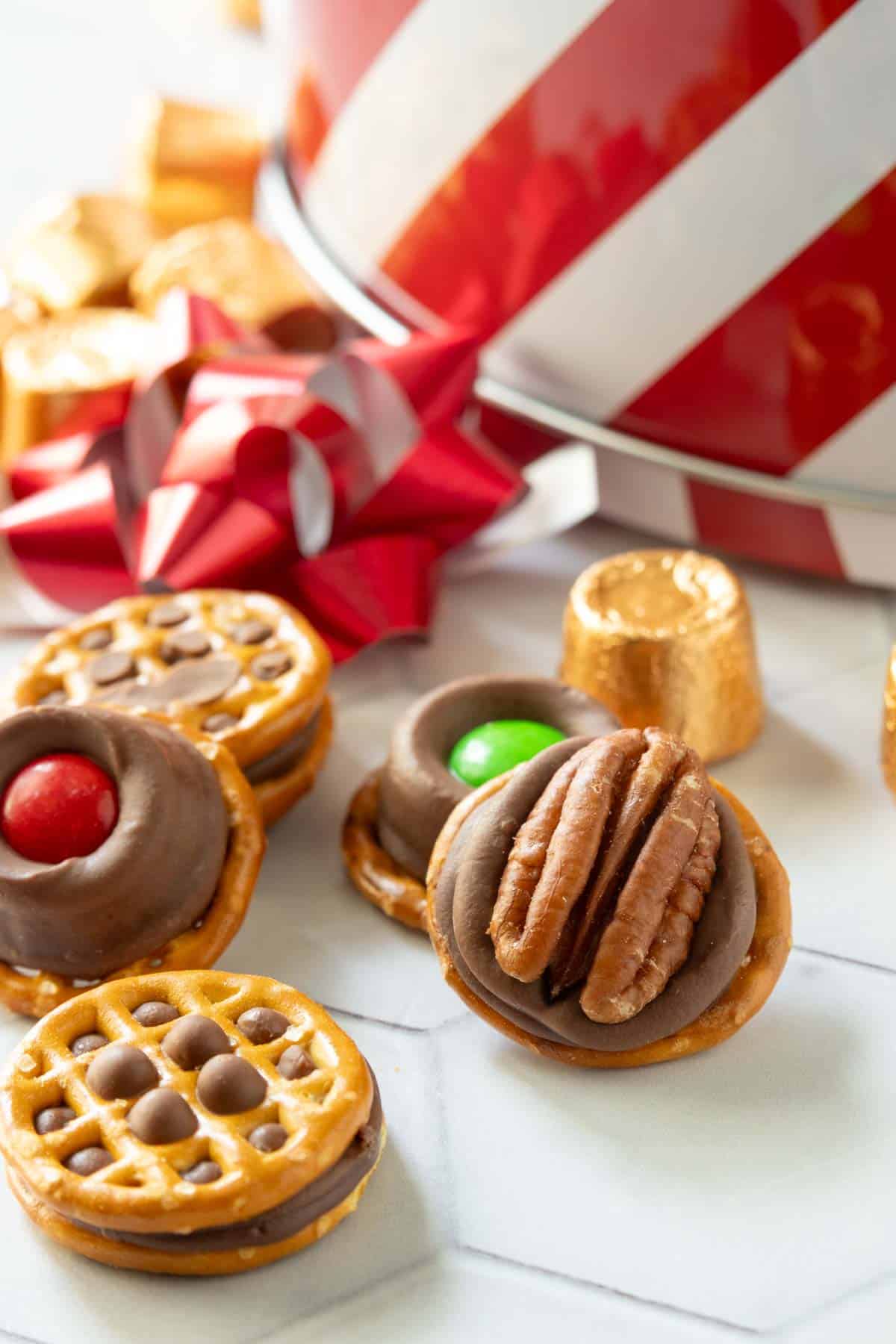 Assorted pretzel bites topped with chocolate, candies, and pecans are arranged on a white surface next to a red and white striped tin and a red bow.