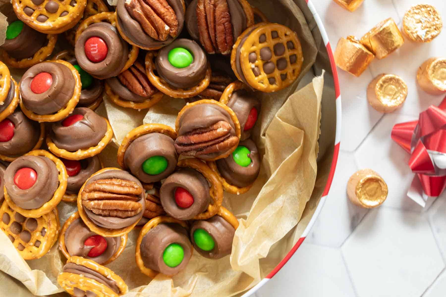 A tin filled with pretzel bites topped with chocolate, pecans, and red or green candies, with additional bites and candies scattered on a white surface nearby.