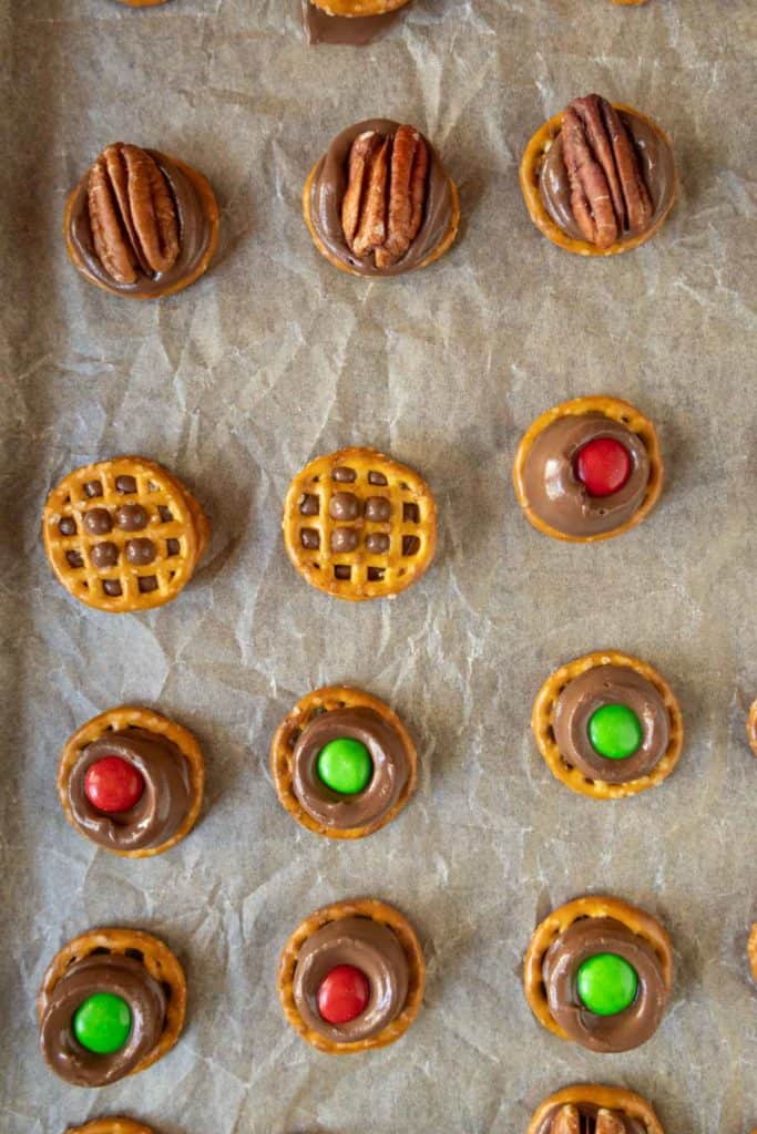Rows of pretzel bites topped with melted chocolate and either a pecan half or a red or green candy, arranged on a parchment-lined baking sheet.