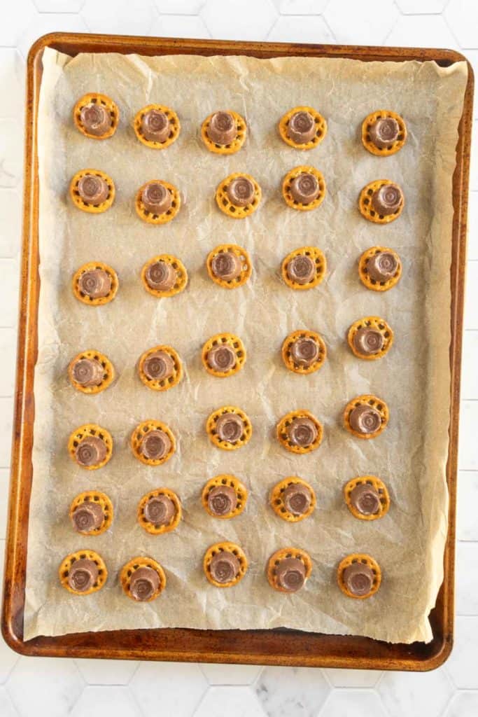 A baking sheet lined with parchment paper holds five rows of pretzel bites topped with chocolate and caramel candies.