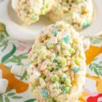 A close-up of a cereal treat shaped like an egg, made with colorful marshmallow cereal pieces, on a floral-patterned surface. Two more treats are on a plate in the background.