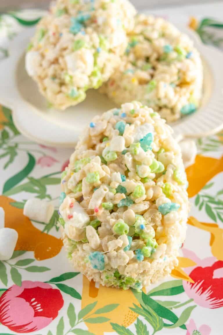 A close-up of a cereal treat shaped like an egg, made with colorful marshmallow cereal pieces, on a floral-patterned surface. Two more treats are on a plate in the background.