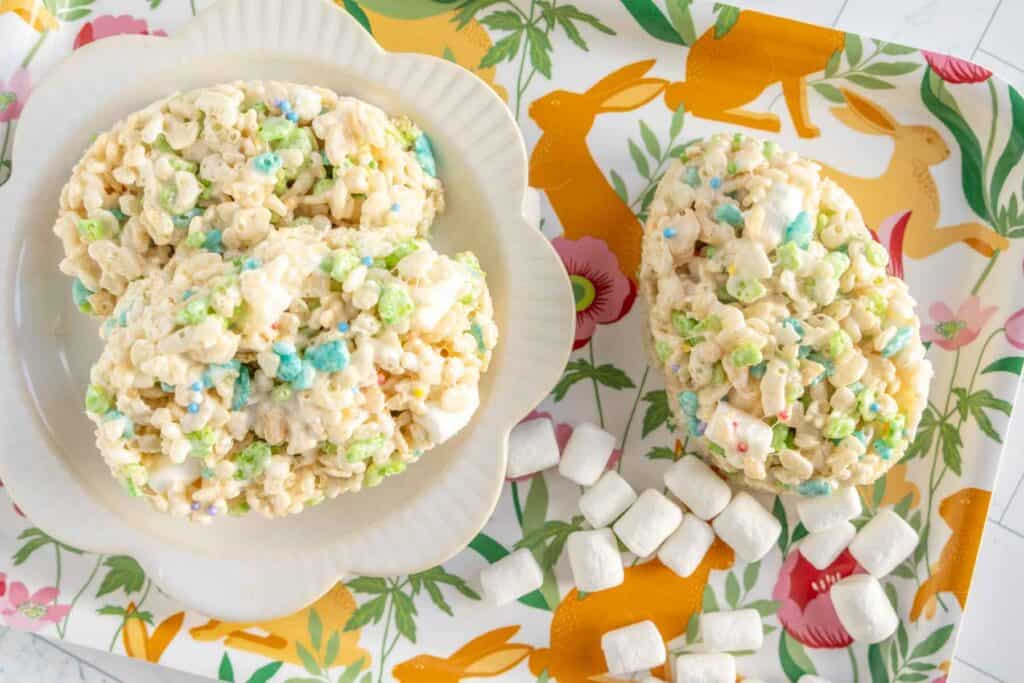 A plate and tray with egg-shaped Rice Krispies treats mixed with colorful marshmallows, surrounded by mini marshmallows, on a decorated tray featuring orange rabbits and flowers.