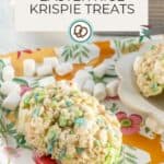 A round Easter rice krispie treat with pastel marshmallows sits on a floral plate, surrounded by mini marshmallows; a baking pan of treats is in the background.