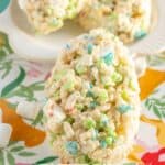 A close-up of Easter rice krispie treats shaped like eggs, made with colorful cereal bits, displayed on a white plate with a floral tablecloth underneath. Text reads "Easter Rice Krispie Treats.