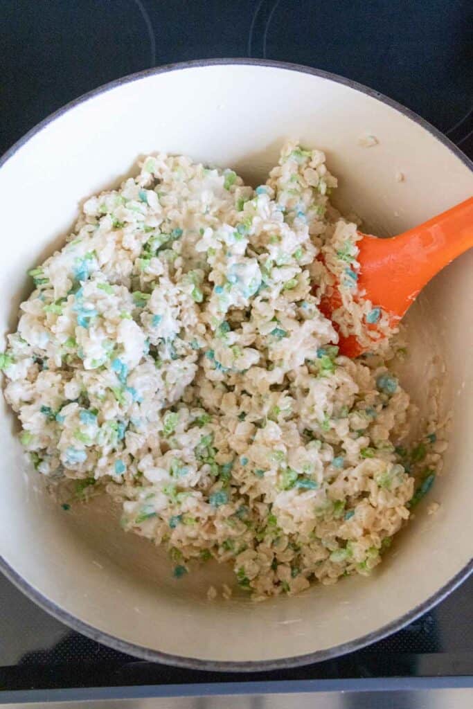 A pot of rice crispy treat mixture with colorful specks being stirred by an orange spatula.
