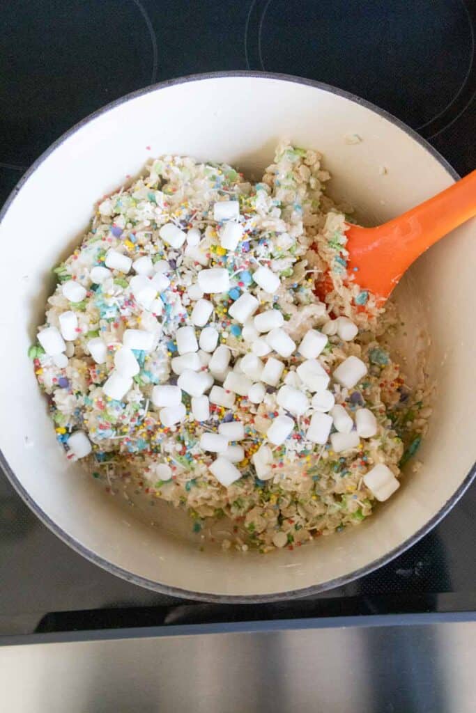 A pot containing rice cereal, marshmallows, and rainbow sprinkles being mixed with an orange spatula.