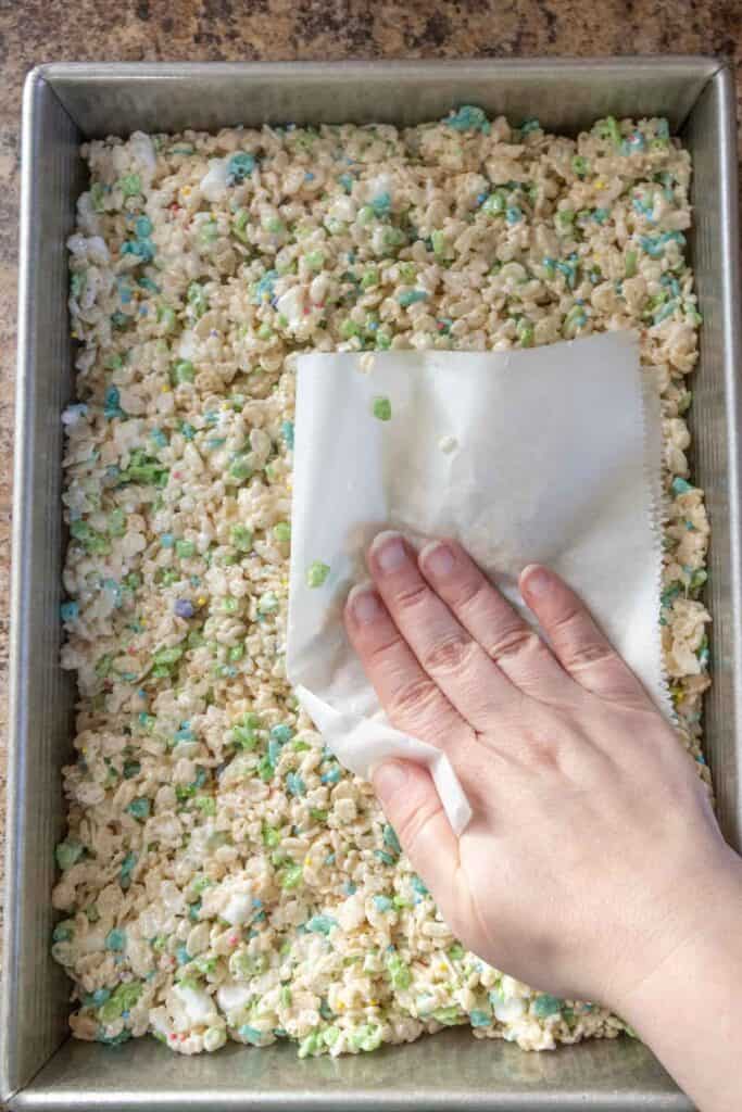 A hand presses Rice Krispies treat mixture with marshmallows and colorful bits into a baking pan using parchment paper.