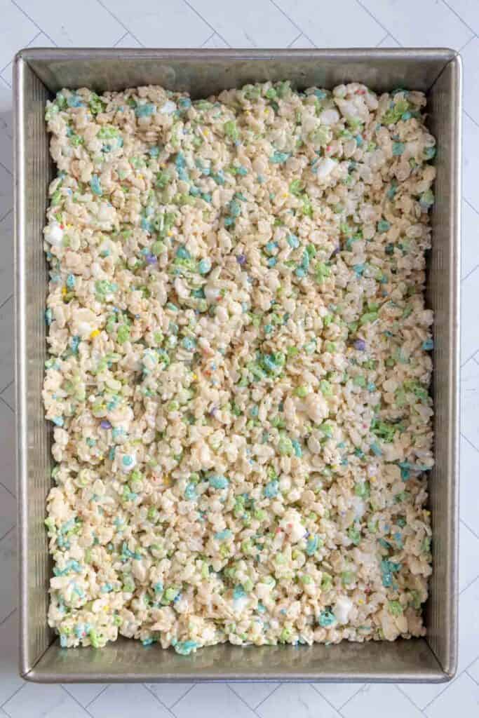 A rectangular metal baking pan filled with a mixture of puffed rice cereal, melted marshmallows, and colorful candy pieces on a white surface.